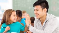 An elementary student and her teacher give each other a high-five at a table in a classroom.