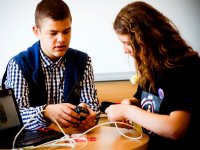 Two teenagers are sitting at a table with a laptop, one is holding a cell phone charger and the other is holding a cell phone, connecting it to a white wire.