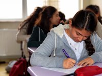 A closeup of a high school girl sitting at a purple desk. She's writing, looking down at her paper. Other students are sitting behind her, slightly blurred.