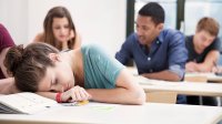 A high school girl is asleep with her head resting on her desk, and other classmates are sitting around her, talking.