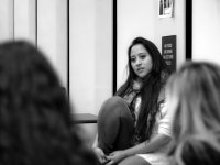 A black and white image of a young, long-haired woman sitting on the floor against a white wall, looking towards two other women sitting on the floor. 