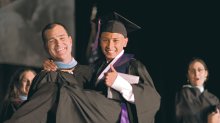 A teacher in a graduation gown is carrying a high school graduate in cap and gown, both are smiling, posing for a photo.