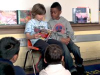 Two young students are sitting in chairs against the wall with a whiteboard behind them. They're reading a book to the rest of the class who are sitting on the carpeted floor in front of them. 