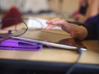 With a blurred background, a student's hand is highlighted hovering over a tablet. 