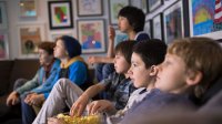 Six preteen boys are sitting on brown couches, eating popcorn, and watching a movie.