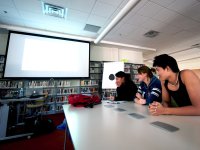 Three high school students are sitting at a long table with a projector in front of them and bookshelves lined across the wall next to them.