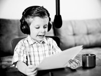 A black and white image of a young boy wearing headphones, reading from papers, speaking into a hanging microphone
