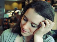 A closeup of a young women in a green shirt resting her head on her hand. The background, a coffee shop, is blurred.