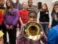 A young girl in her preteens is sitting, staring at the camera, playing a trumpet. She's in a classroom, surrounded by eight other students and her teacher who are all slightly out of focus.