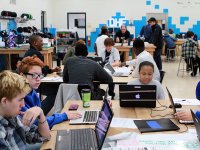 A classroom filled with high school students sitting at large rectangular tables, working together on laptops. 