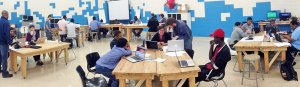 A panoramic view of a large classroom. The walls are painted in white and blue blocks, like a digital screen. Five groups of students are sitting at sets of rectangular, wood tables pushed together. They're all working on laptops.