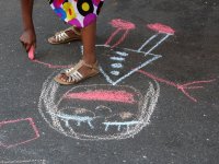 A girl in golden sandals and a colorful polka dot dress is standing on cement, bending over, holding pink chalk, drawing a smiling girl.