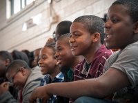 A closeup of a large group of young, black kids smiling, sitting in bleachers in a multipurpose room