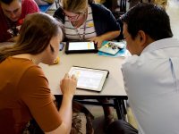 Four teachers are sitting around a table, looking at spreadsheets together on two tablets