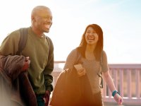 Two young adults wearing backpacks are walking outside, laughing, with jackets in their hands.