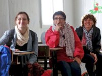 Three women are sitting at desks, looking at the front of the classroom, smiling.