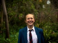 A young, teenage girl in a white collared shirt, blue and red checkered tie, and a blue jacket is standing, smiling, with trees as her backdrop. 