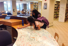 A photo of 2 female students writing a whiteboard table with colored markers.