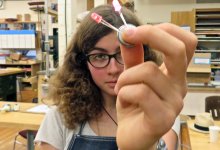 A curly-haired teenage girl is standing in a classroom holding a metal circle that's connected to two miniature red light bulbs. One of them is lit. 