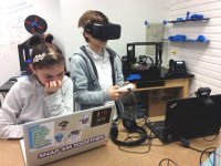 A young girl is sitting at a classroom table working on her laptop. A young boy is standing next to her with high-tech eye goggles on, a video game controller in his hands, and he's looking at a laptop with a video camera connected on the top. 