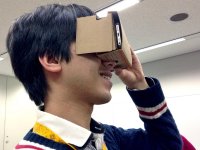 A teenage boy is holding a Google Cardboard viewer up to his eyes, a cardboard box with his smart phone inside of it, looking into it as if it were binoculars or a camera. 