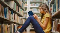 A teenage girl sits on floor and reads book in library. 