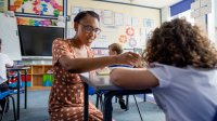 Elementary school teacher kneels down next to a students desk to help her