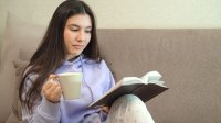 Teen girl reads a book while sitting on a couch and drinking from a mug