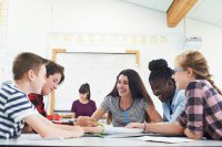 A group of five students working together in a classroom, their teacher sitting at a desk doing paperwork in the background