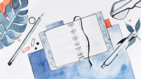 A watercolor illustration of a journal laying open on a desk, surrounded by various objects such as pens, glasses, pencils, and paperclips