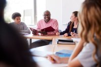 A counselor sitting at a circle of desks with high school students, holding an open binder