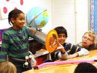 Boy showing his project to classmates -- a silver tube over his arm with a globe tied to the top and a fan at the end of the tube