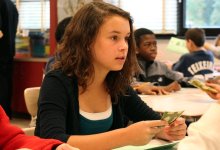 A young girl is sitting in class, looking straight ahead with a playing card in her hand and a green sheet of paper on her desk. Other students in the classroom are sitting with green sheets of paper and playing cards about. 