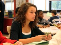 A young girl is sitting in class, looking straight ahead with a playing card in her hand and a green sheet of paper on her desk. Other students in the classroom are sitting with green sheets of paper and playing cards about.