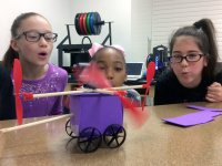 Three upper elementary girls are squatting next to a table, blowing on the wings of a toy car they've made, propelling it to move. 