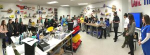 Panoramic shot of students standing around work tables listening as four adults speak. There are projects on the walls, and lots of tools, bins, and notebooks all over the tables.