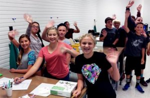 A dozen students working with pens and paper waving to the camera