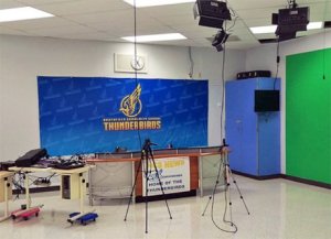 A recording studio with the school logo as a backdrop behind the news desk, overhead lights, a mix board, and a wall with green screen