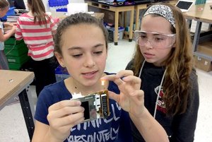 A girl is holding a student-made electronic device that has lit a small light bulb. Another girl in protective goggles is standing next to her.