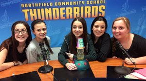 Five girls are sitting at a table with microphones behind a backdrop that says, 'Northfield Community School Thunderbirds.'