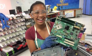 A girl wearing safety goggles, apron, and gloves, smiling holding up a computer circuit board