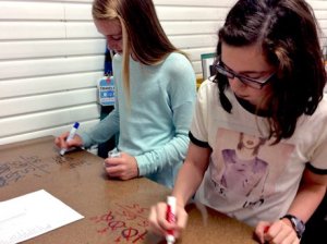 Two girls at one large desk holding dry erase markers writing math equations directly on the desktop