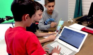 Three boys working together; one using an app on a laptop, two others working with pipe cleaners and paper