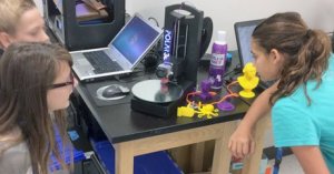 Three girls around a table watching something being printed in 3D