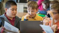 A closeup of three young boys sitting at a classroom table looking at an opened laptop together. 