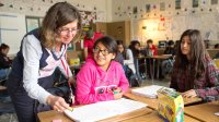 A female middle school teacher talks with two female students who are seated at their desks.