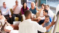 A group of teachers sit in a discussion circle while one stands pointing at a white board.