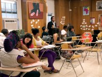 A group of students are sitting at their desks in class, listening to their teacher speak.