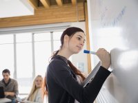 A high school female is holding a notebook in one hand and writing on a whiteboard at the front of the class with another hand. Two other students are sitting at a table nearby her.