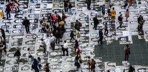 People walking through the square that has the ground wall papered with black and white photo headshots of hundreds of people